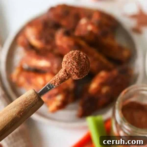 A close up shot of buffalo chicken dry rub in a measuring spoon above a plate of buffalo chicken wings.