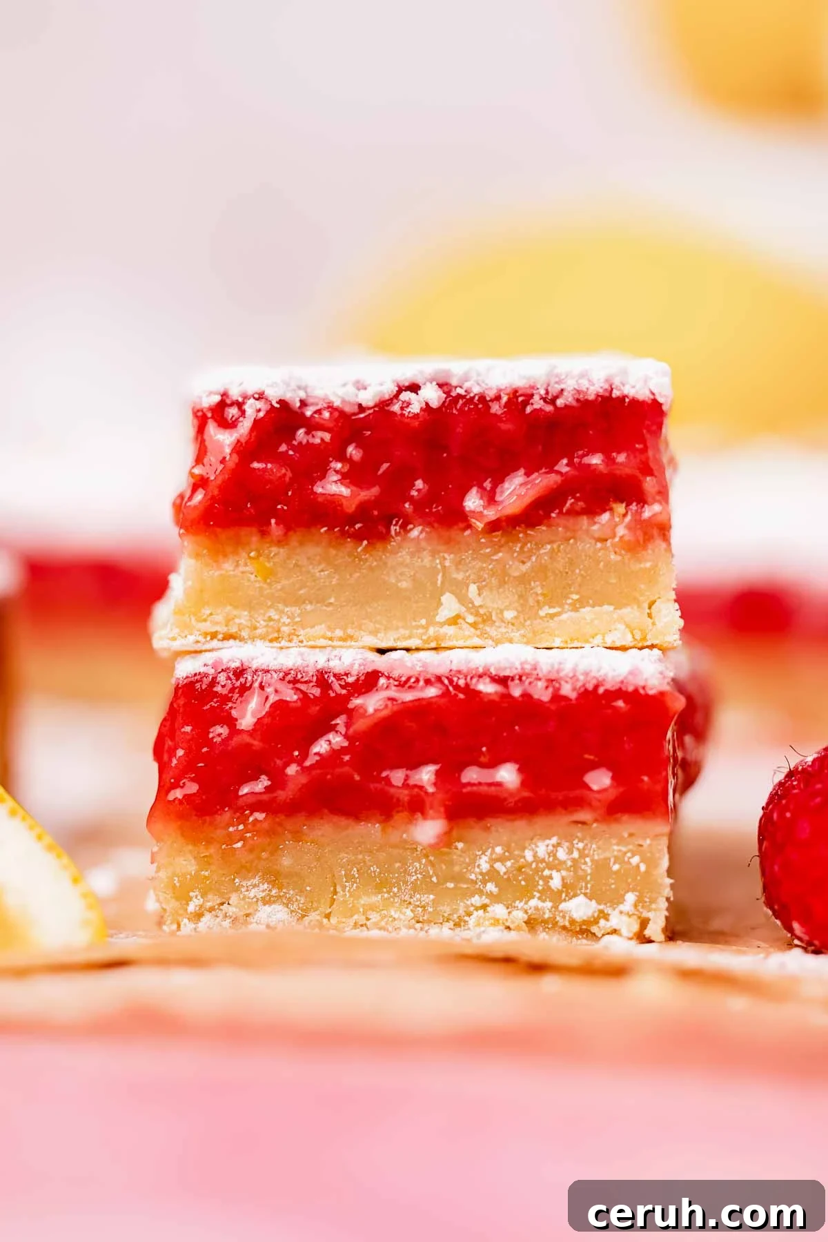 two stacked lemon raspberry bars on parchment paper with fresh lemons and raspberries surrounding