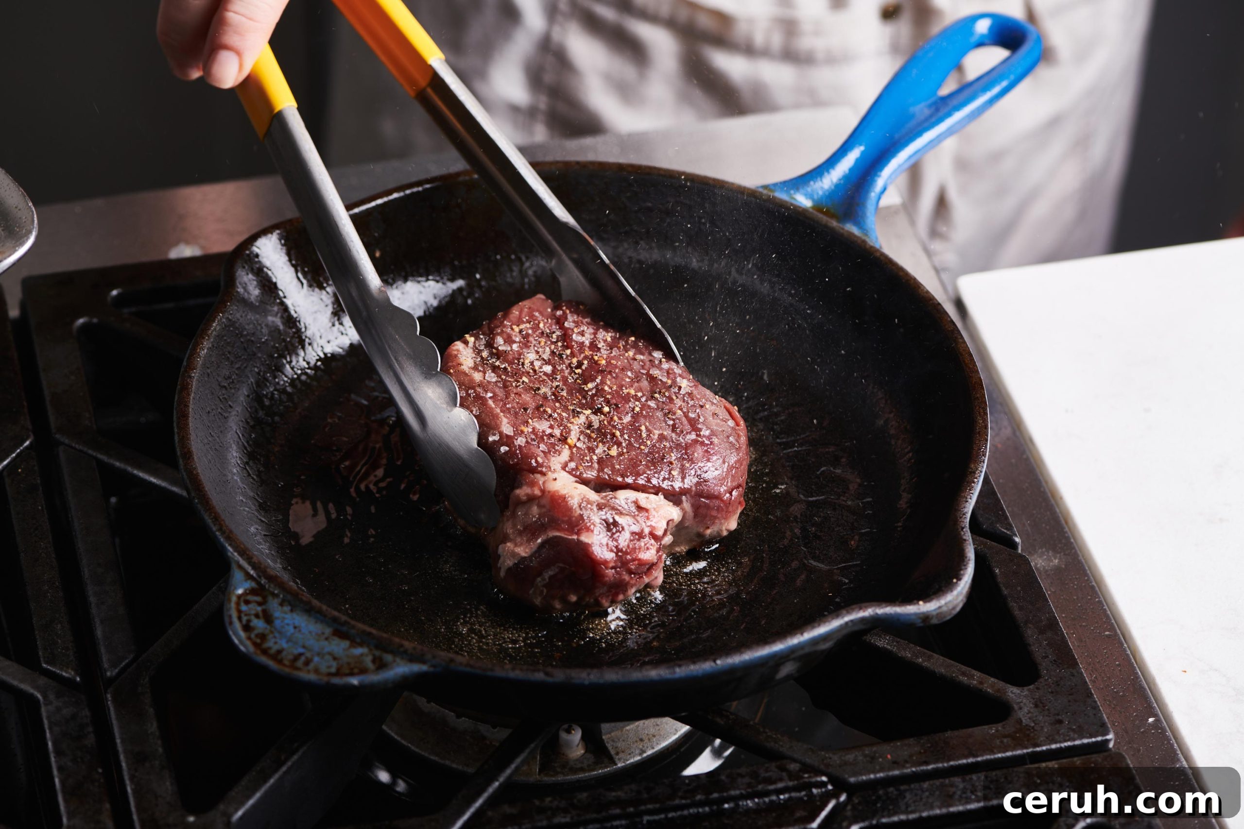 Tongs grabbing a Filet Mignon in a cast iron skillet.