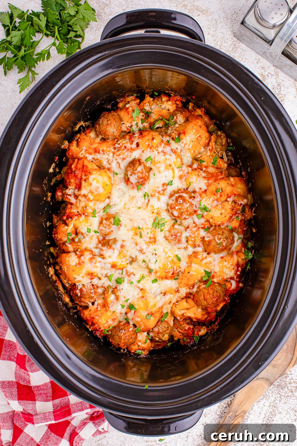 A slow cooker containing meatballs in a tomato sauce, topped with melted cheese and garnished with chopped herbs.