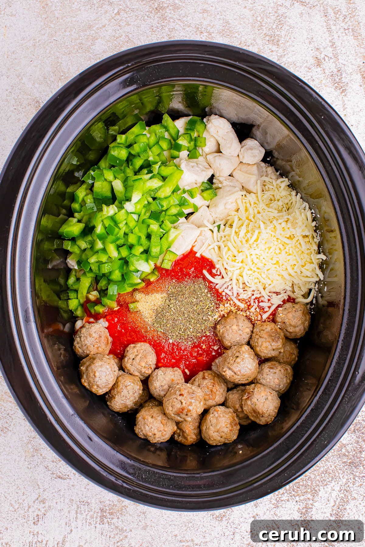 A slow cooker filled with chopped green peppers, diced chicken, shredded cheese, tomato sauce, pepper, and meatballs, ready for cooking.