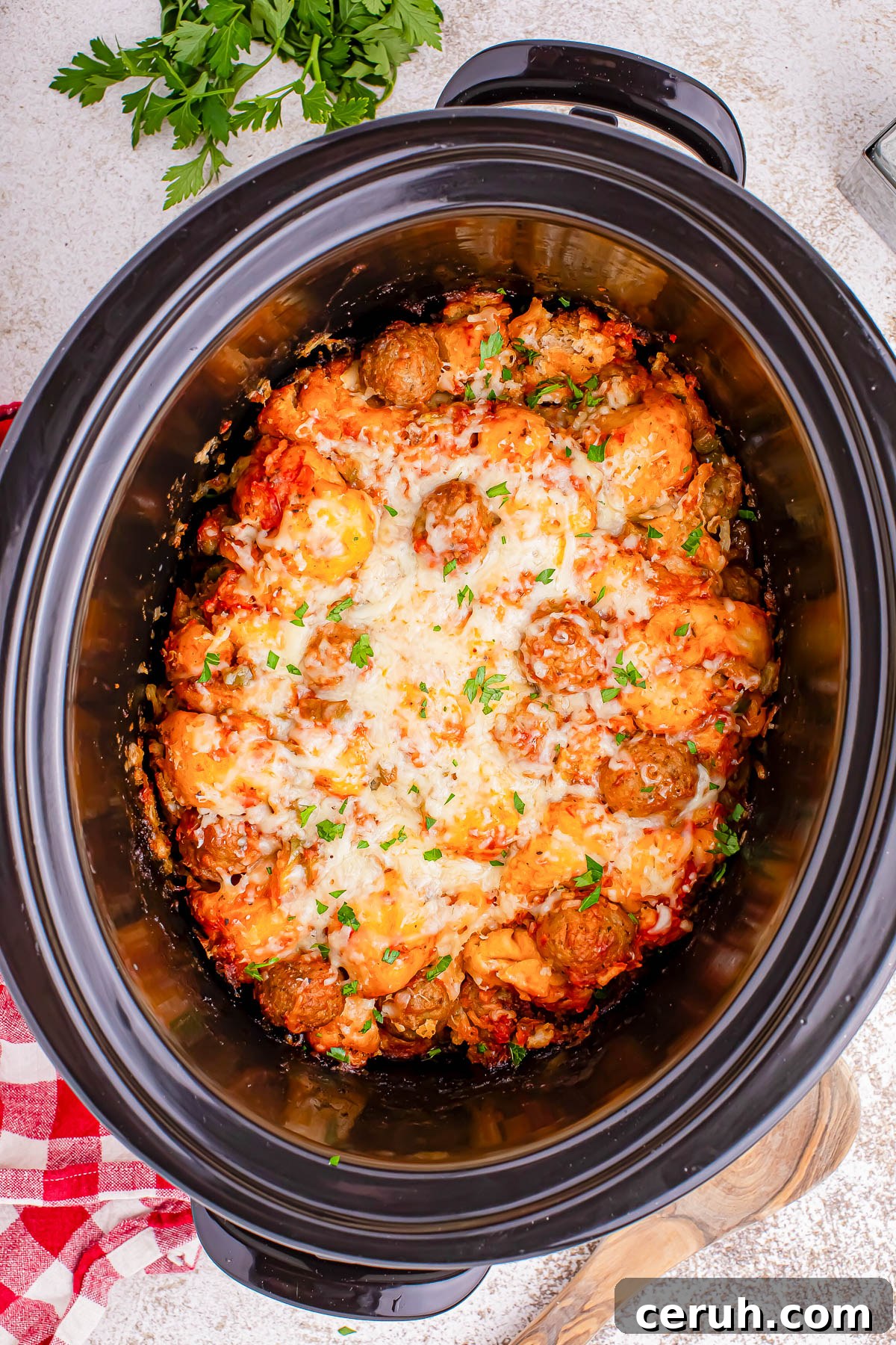 A slow cooker filled with a cheesy dish, including meatballs and pasta, garnished with herbs. A red and white cloth and some parsley are placed nearby.