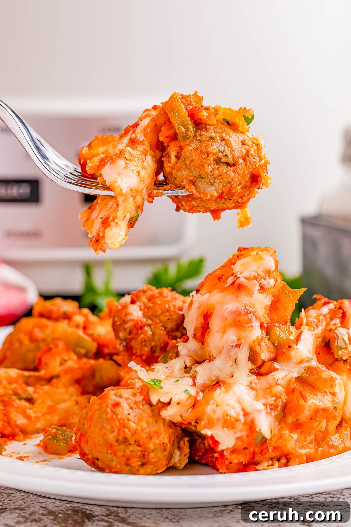 A fork lifts cheesy meatballs with tomato sauce above a plate filled with more meatballs and melted cheese.