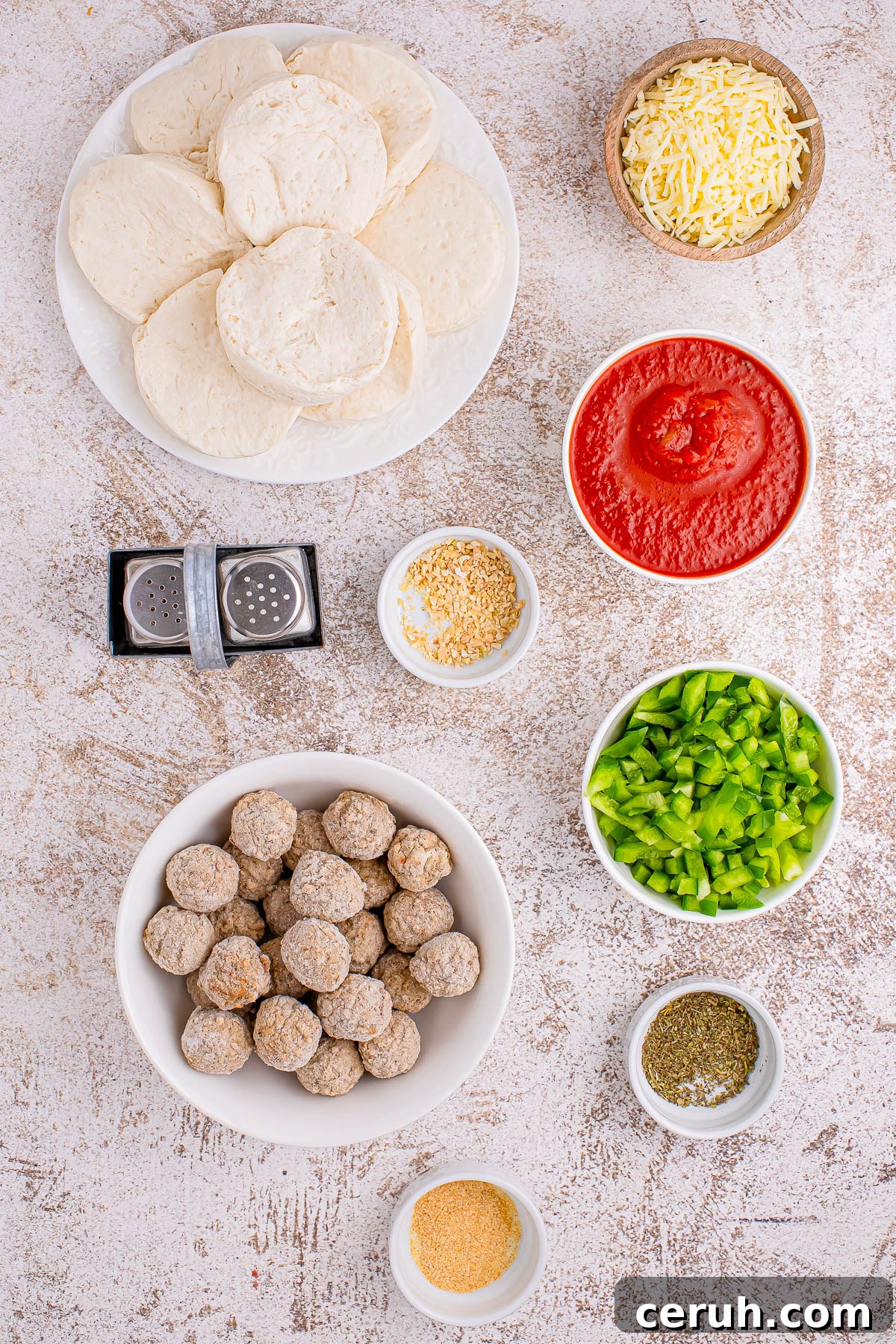 Ingredients arranged for pizza assembly, including biscuit dough, grated cheese, tomato sauce, sliced green peppers, meatballs, and seasoning bowls on a textured surface.