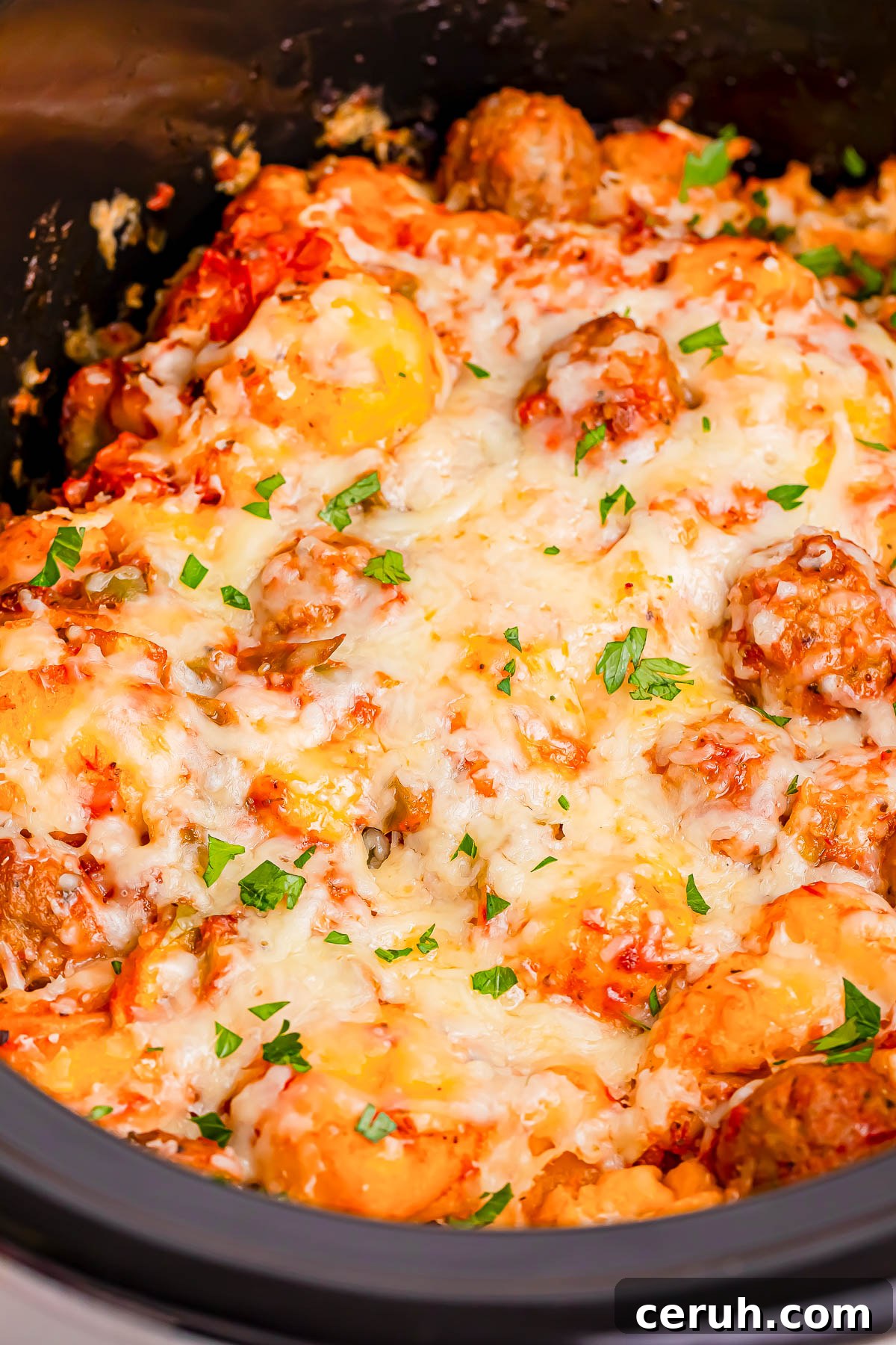 Close-up of a cheesy baked dish with meatballs and herbs, topped with melted cheese and garnished with chopped parsley.
