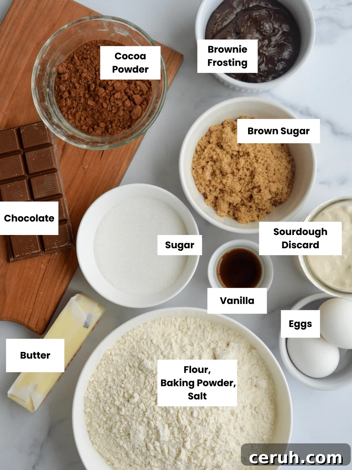 Ingredients are in small containers on a countertop and include brownie frosting, cocoa powder, brown sugar, chocolate, sugar, sourdough discard, vanilla, eggs, butter, flour, baking powder, and salt.