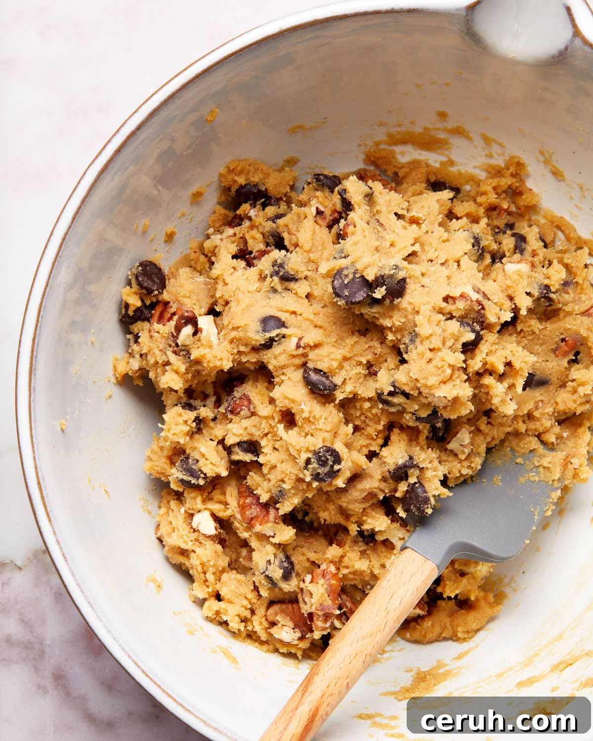 Chocolate Chip and Pecan Cookies — Chewy Oven-Baked Recipe 4 Cookie dough in a white bowl with a spatula inserted into it.