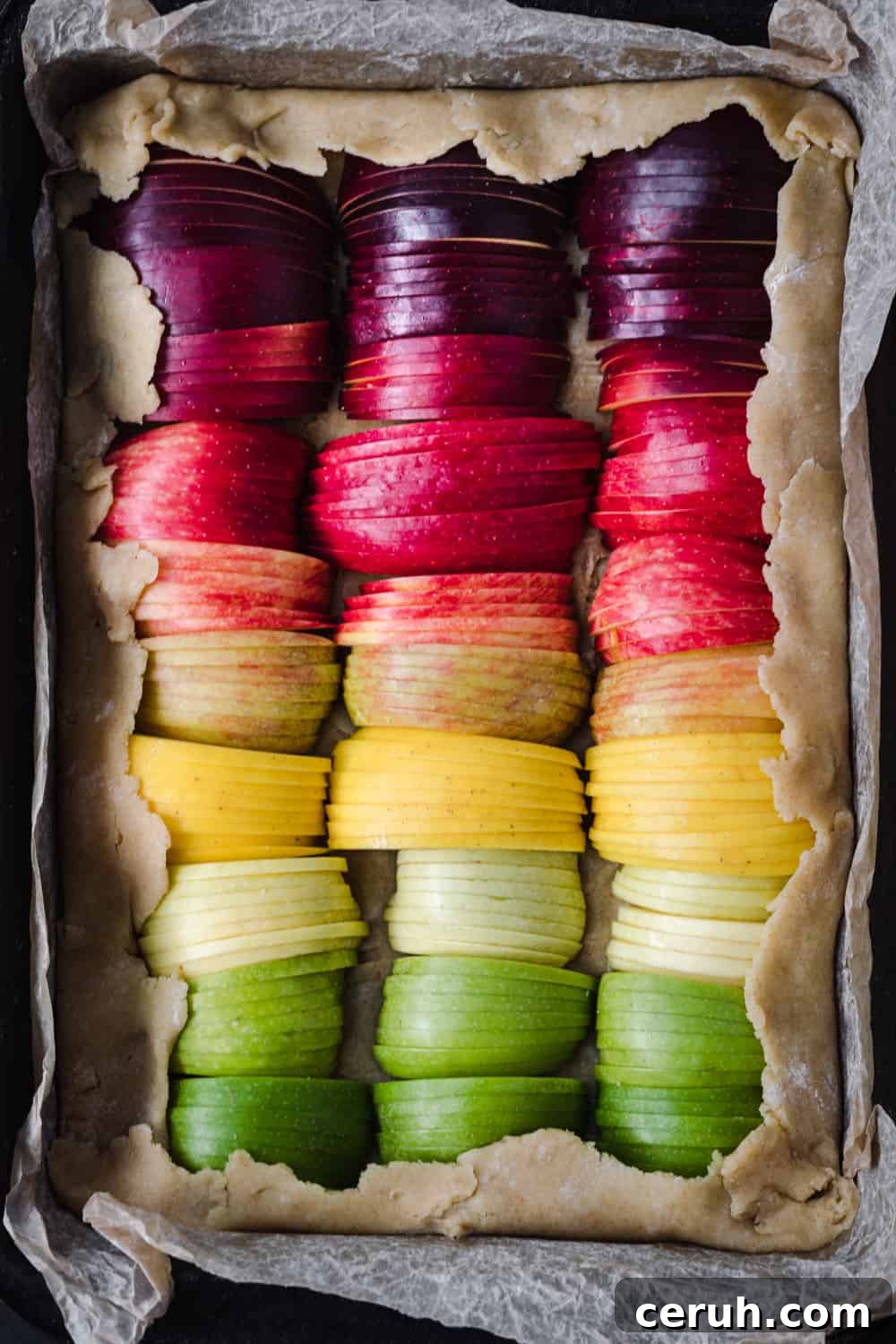 Thinly sliced apples for an apple galette, arranged like a rainbow on galette dough in a baking tray with parchment paper; overhead.