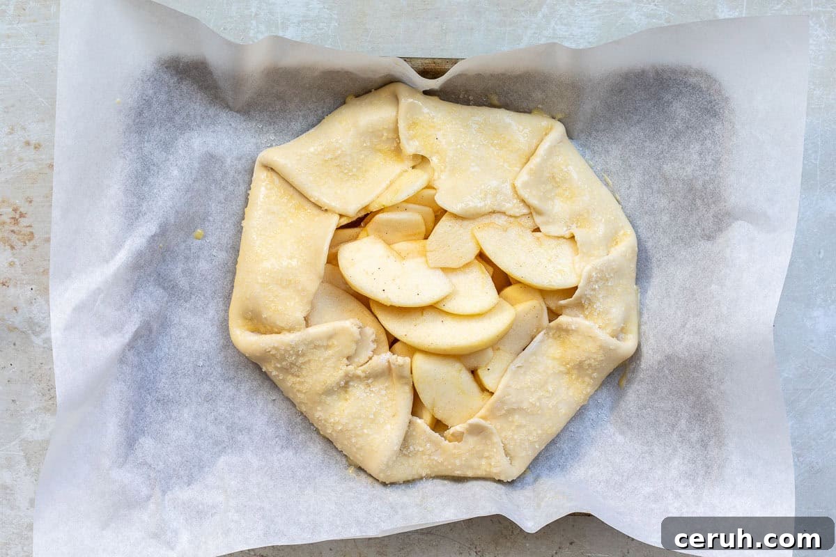 A rustic, unbaked apple galette with folded edges, filled with sliced apples on parchment paper. The crust is sprinkled with sugar, and the galette is on a baking tray ready to be baked.
