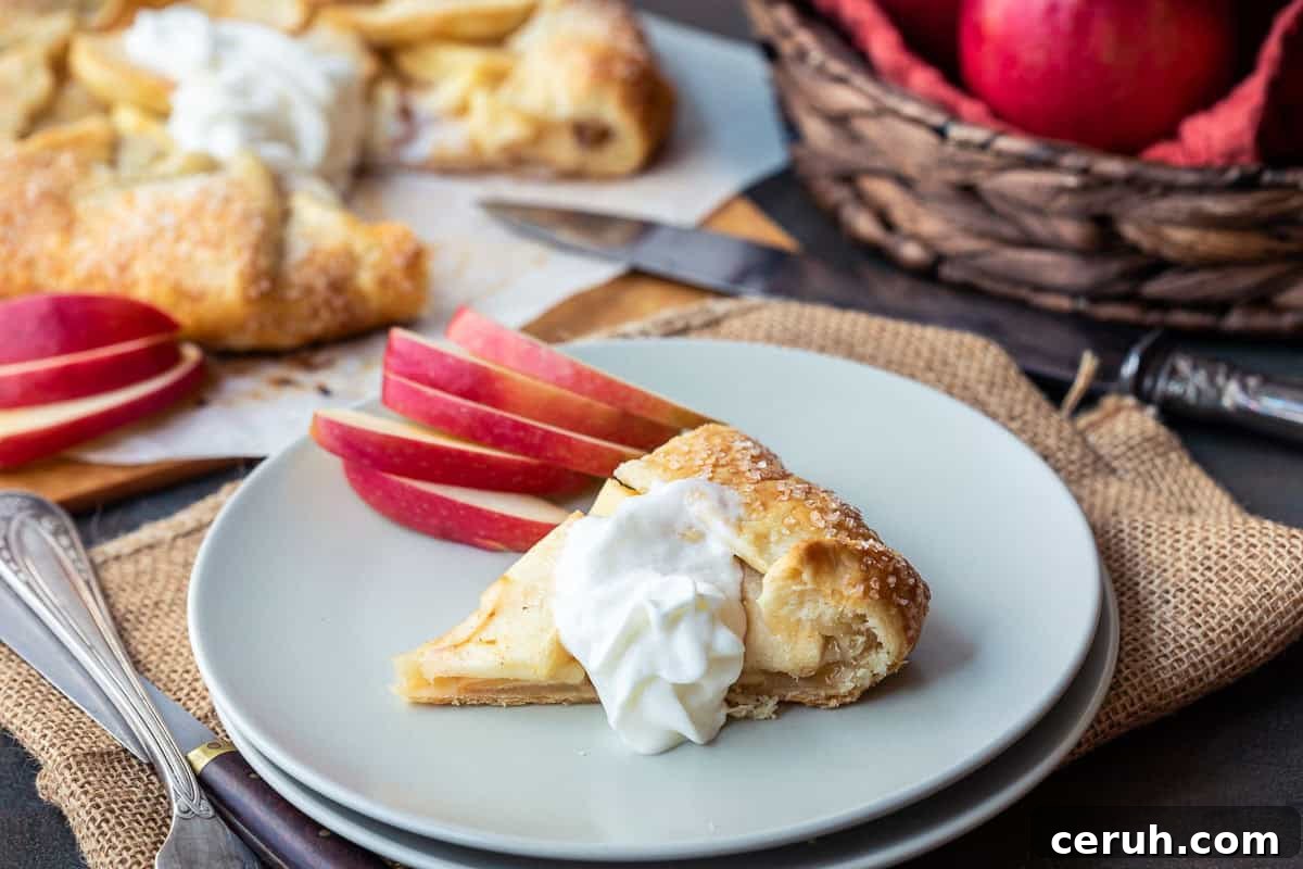 A slice of apple galette topped with whipped cream on a plate, with sliced red apples beside it. In the background, there's more tart on parchment paper and a basket of red apples. A knife rests nearby on a burlap cloth.