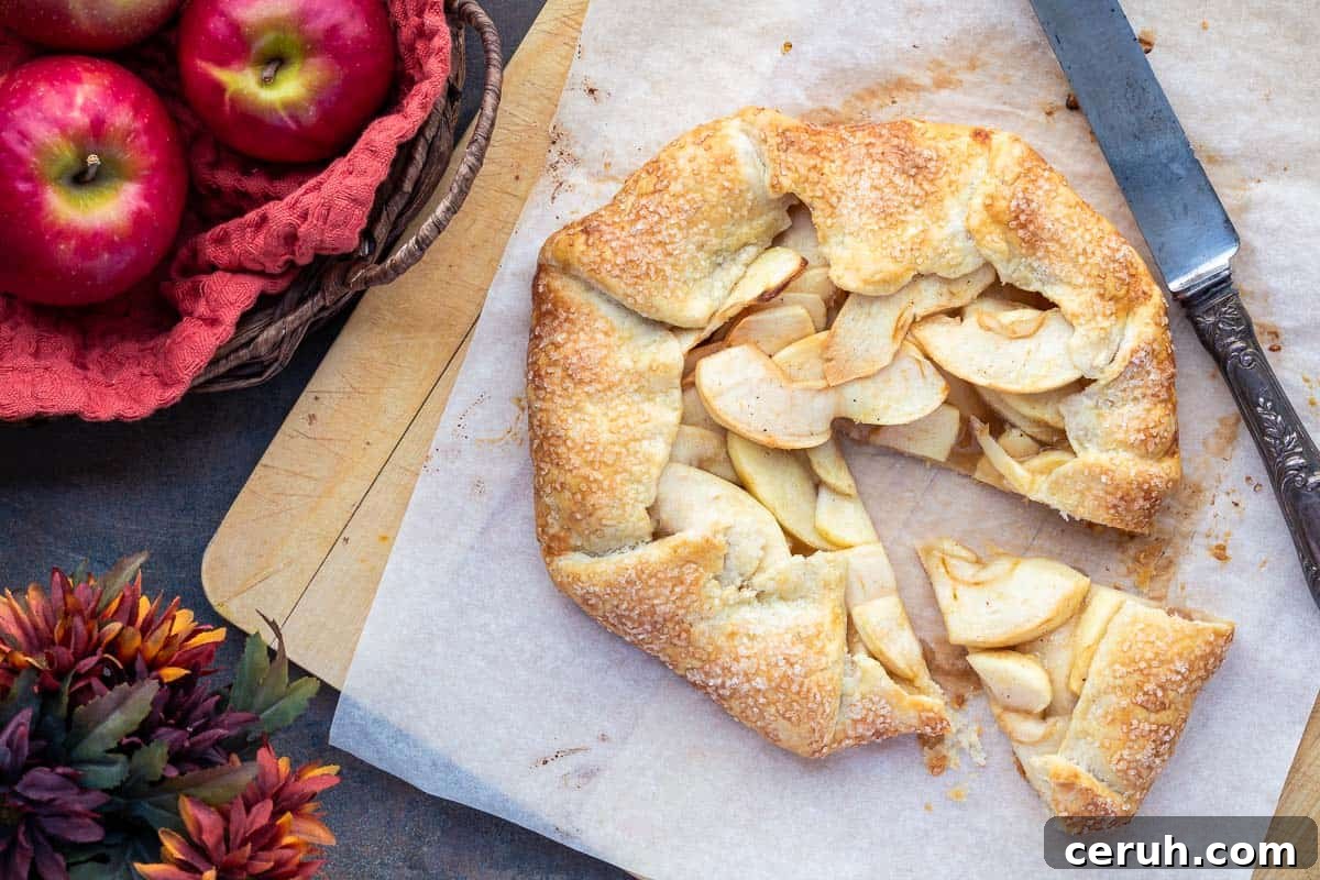 A rustic apple galette on parchment paper, partially sliced, with a vintage knife nearby. A basket of red apples and a cloth is on the left, along with autumn flowers in the corner.
