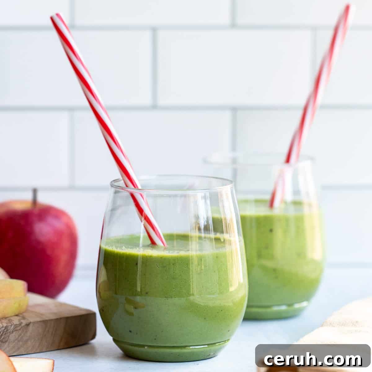 apple spinach smoothie with red and white straw next to 2nd smoothie glass, cutting board, and apples.