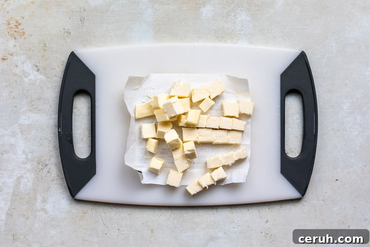Diced cubes of butter on a sheet of parchment paper placed on a white cutting board with black handles, viewed from above. The surrounding surface is a light, textured countertop.