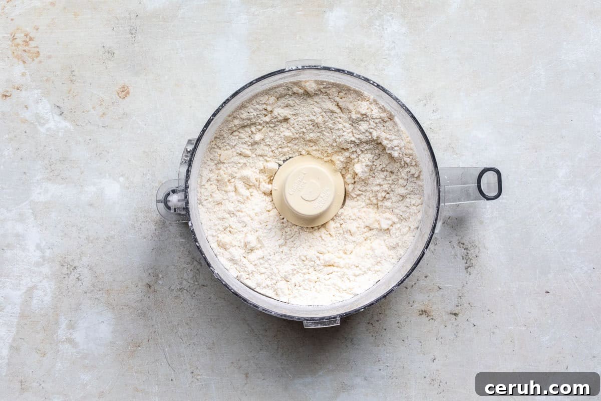 A food processor bowl filled with a flour/butter mixture, sitting on a textured, light-colored surface. The processor lid is off, showing a central plastic blade.