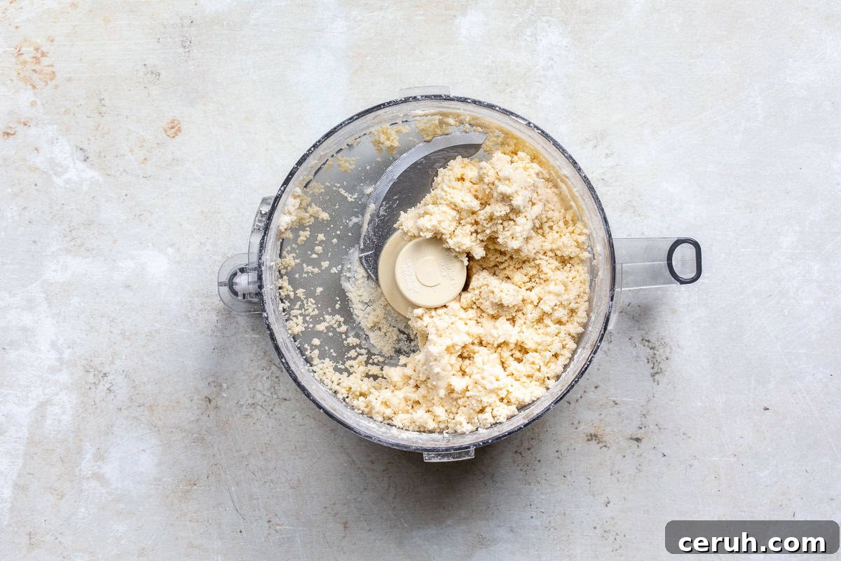 A food processor bowl filled with crumbly pale dough on a light, textured surface. The pie dough is rough and unevenly distributed around the central blade.