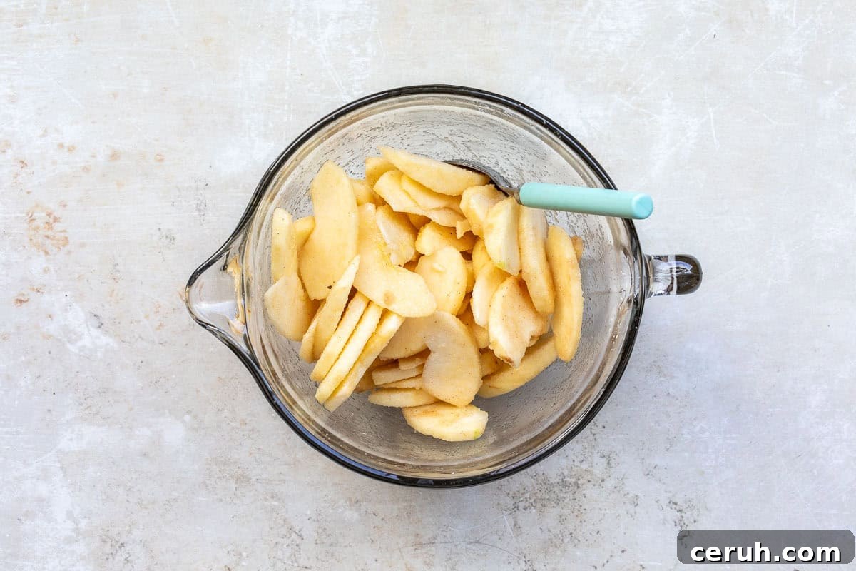 A glass mixing bowl filled with sliced apples, lightly coated, with a teal spoon resting inside. The background is a textured light surface.