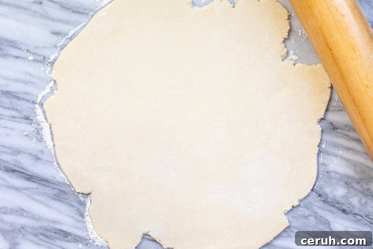 A rough circle of rolled-out galette dough on a gray and white marble surface, with a wooden rolling pin resting on the right side.