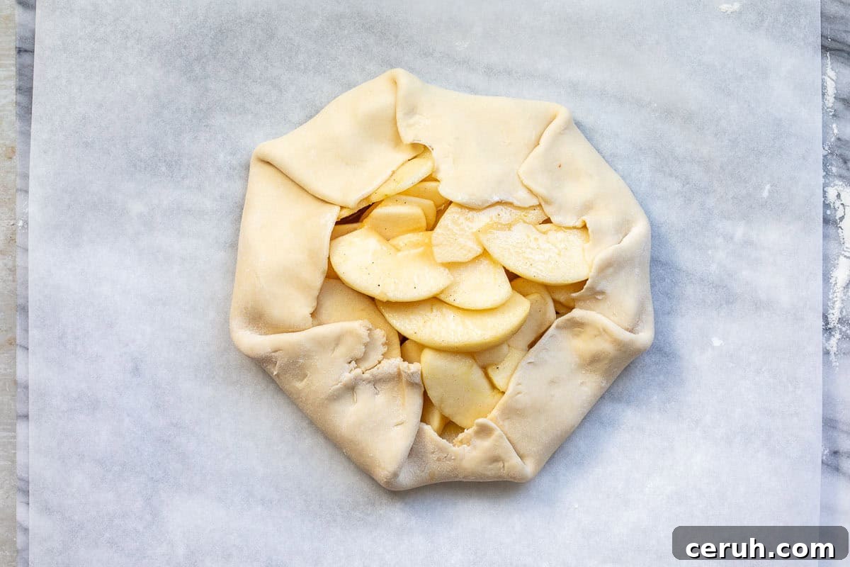 A partially folded apple galette on parchment paper, showing slices of apples stacked in the center of a round pie crust with folded edges. The galette is ready to be baked.
