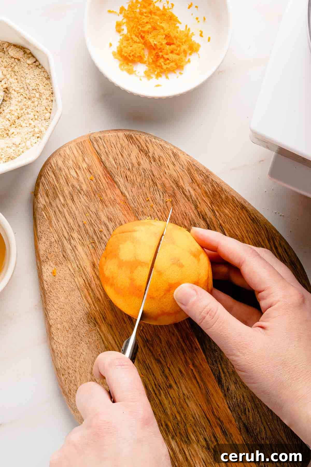 zested orange being cut in half on wooden cutting board.