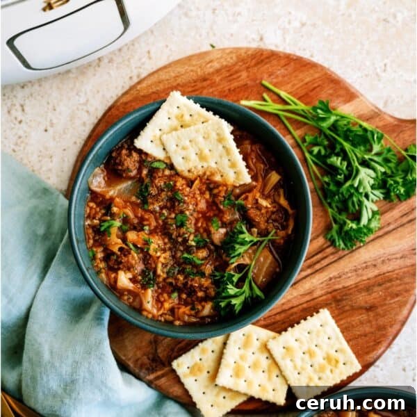 A bowl of cabbage roll soup garnished with parsley and served with saltine crackers, on a wooden board with fresh parsley and a slow cooker in the background.