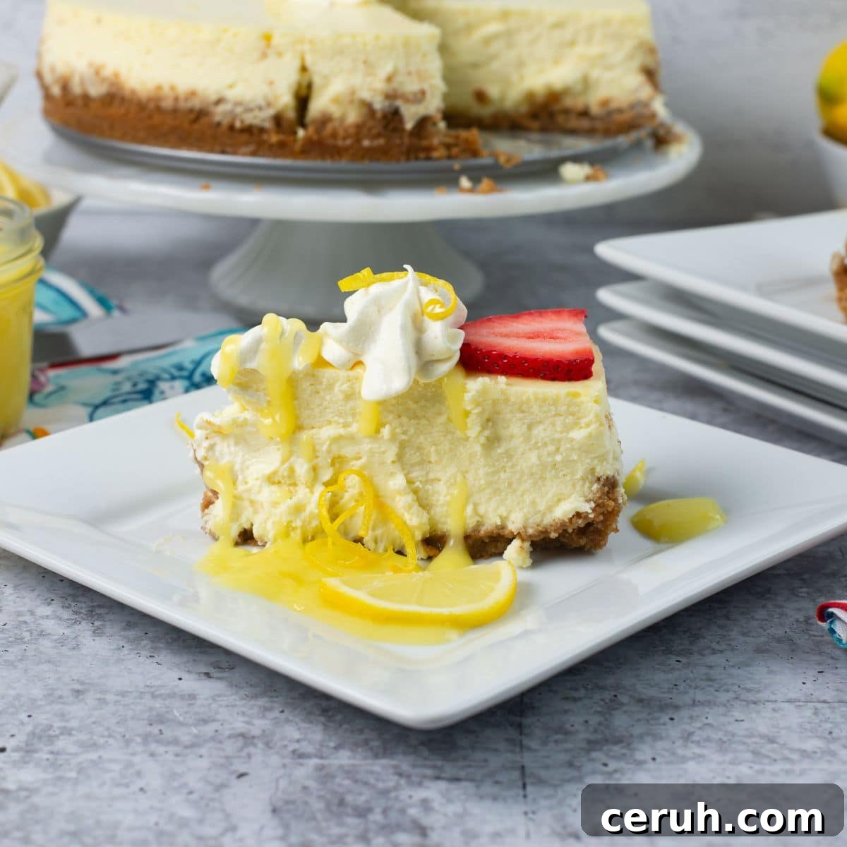 A slice of Lemon Cheesecake on a plate garnished with whipped cream, lemon curd, and a slice of strawberry.