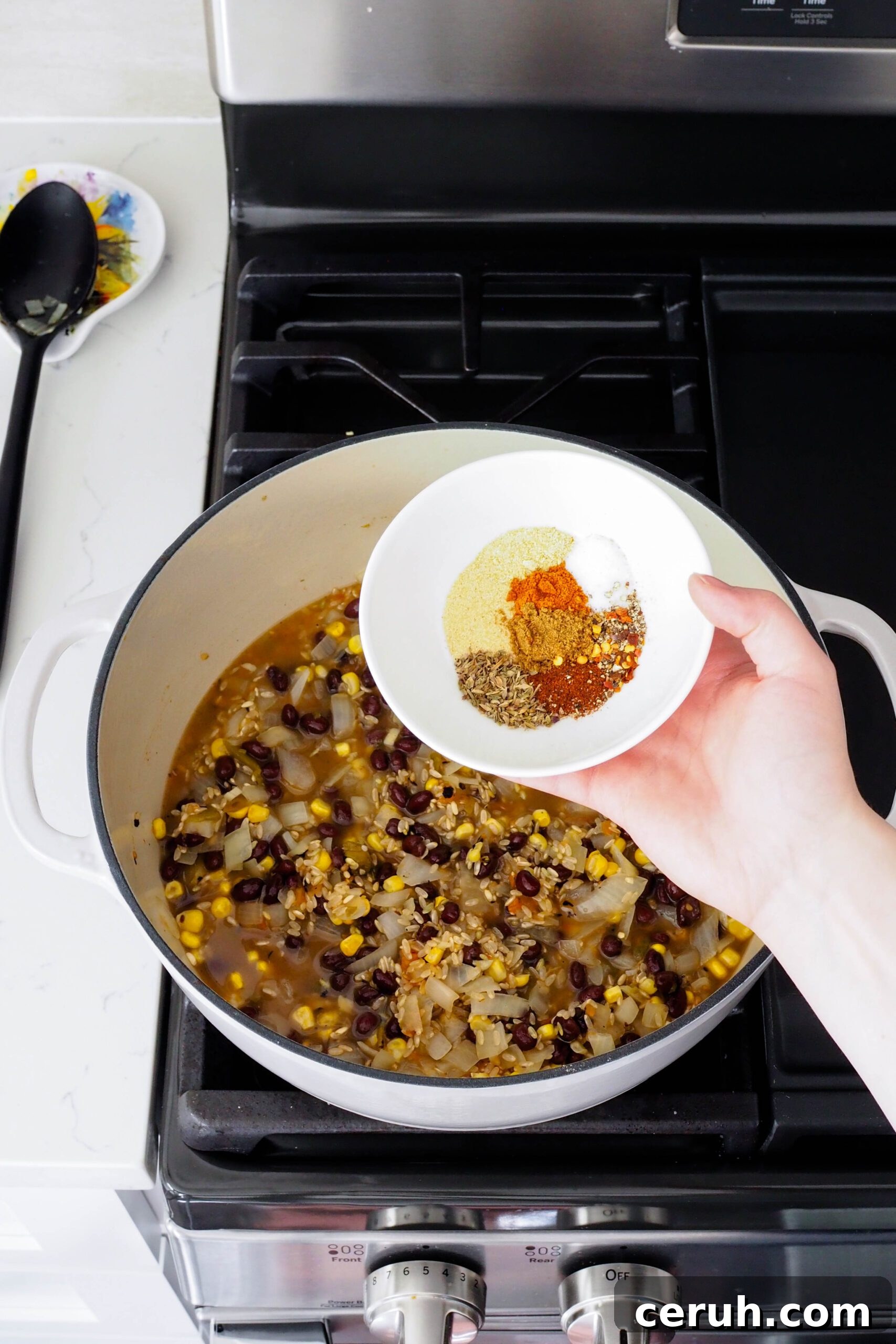 A small bowl of spices is held over a full Dutch oven.