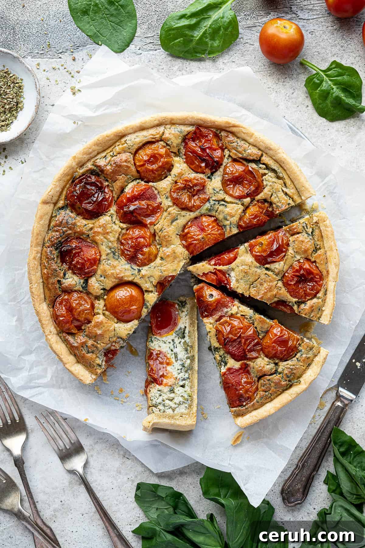 The sliced vegan tomato spinach quiche on a sheet of baking parchment surrounded by fresh basil, spinach and tomatoes.