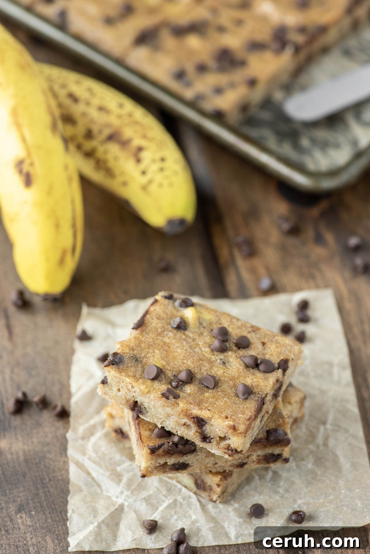 3 banana bars on parchment paper with baking sheet and bananas in background