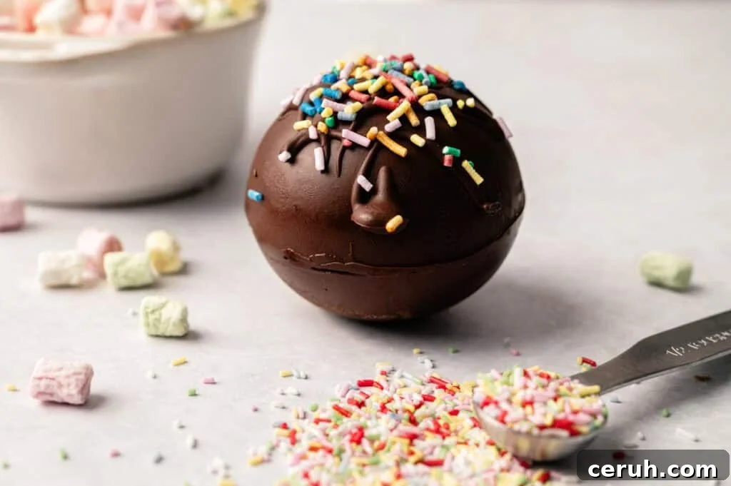 A Hot Chocolate Bomb covered with colorful sprinkles sits on a white surface, surrounded by loose sprinkles, mini marshmallows, and a measuring spoon.