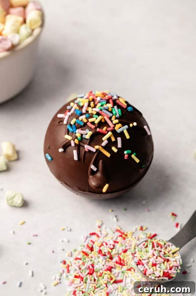 A round Hot Chocolate Bomb topped with colorful sprinkles sits on a light surface, surrounded by extra sprinkles and mini marshmallows, ready to create a deliciously sweet treat.