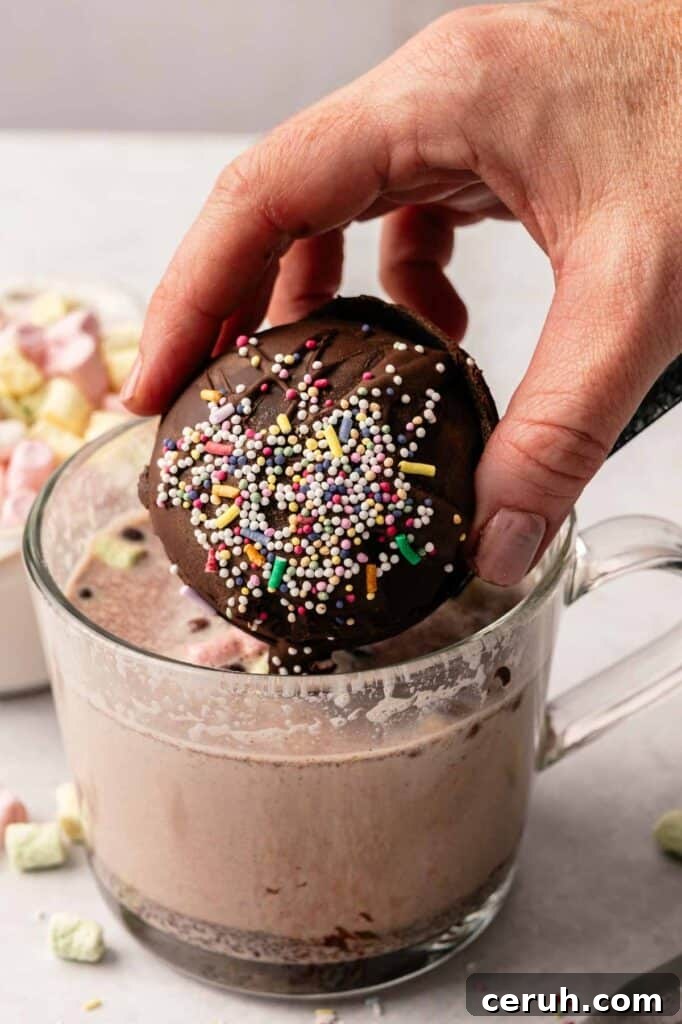 A hand dips a chocolate-covered Hot Chocolate Bomb with colorful sprinkles into a glass mug of hot chocolate; marshmallows are visible in the background.