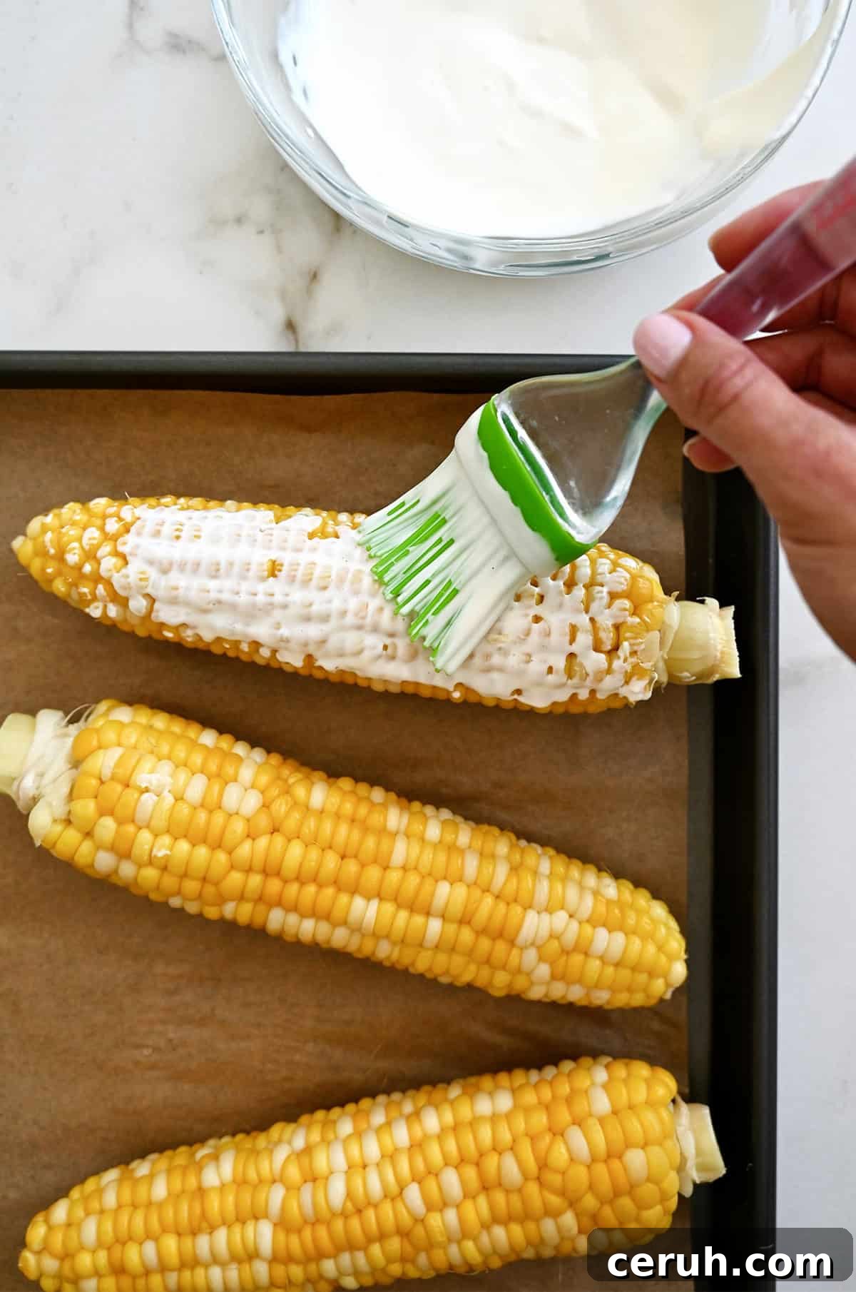 Using a silicone pastry brush to slather boiled corn cobs with a creamy sauce.