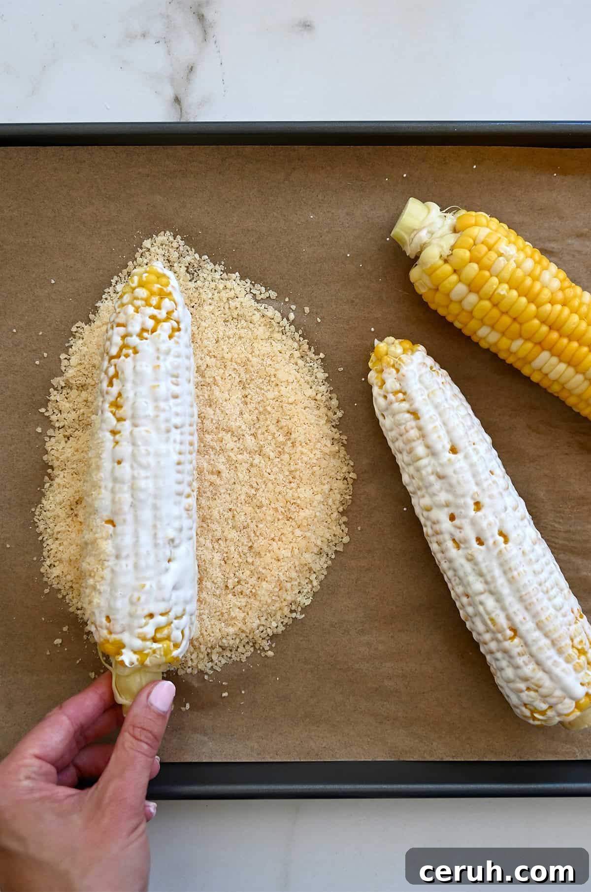 Hand rolling a freshly cooked ear of corn, coated in creamy elote sauce, through crumbled Cotija cheese for Mexican street corn.