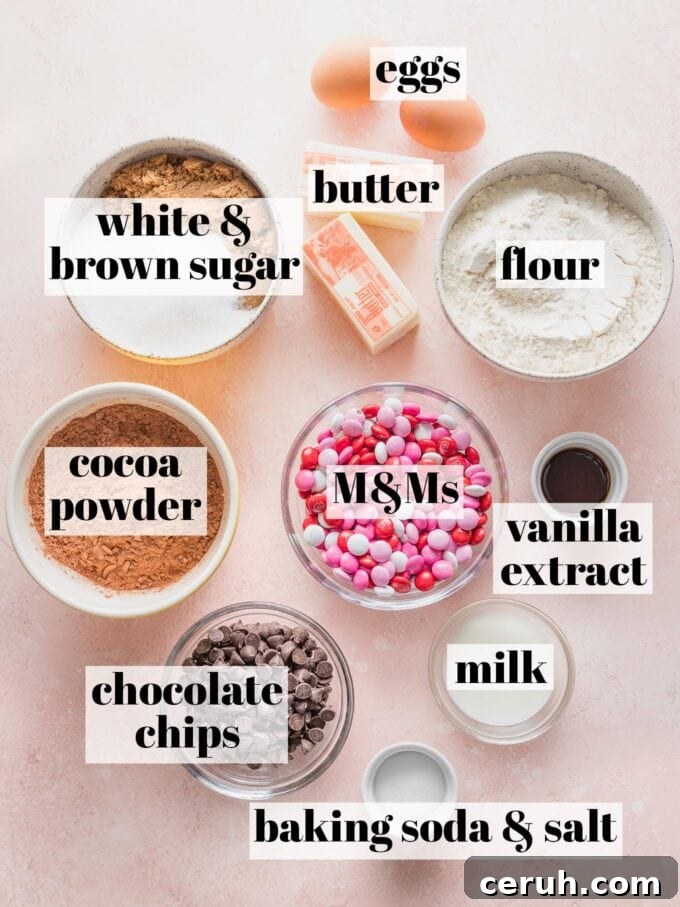 Labeled overhead image of butter, flour, sugar, cocoa powder, vanilla extract, eggs, milk, baking soda, salt, chocolate chips, and M&Ms all in prep bowls and ready to bake.