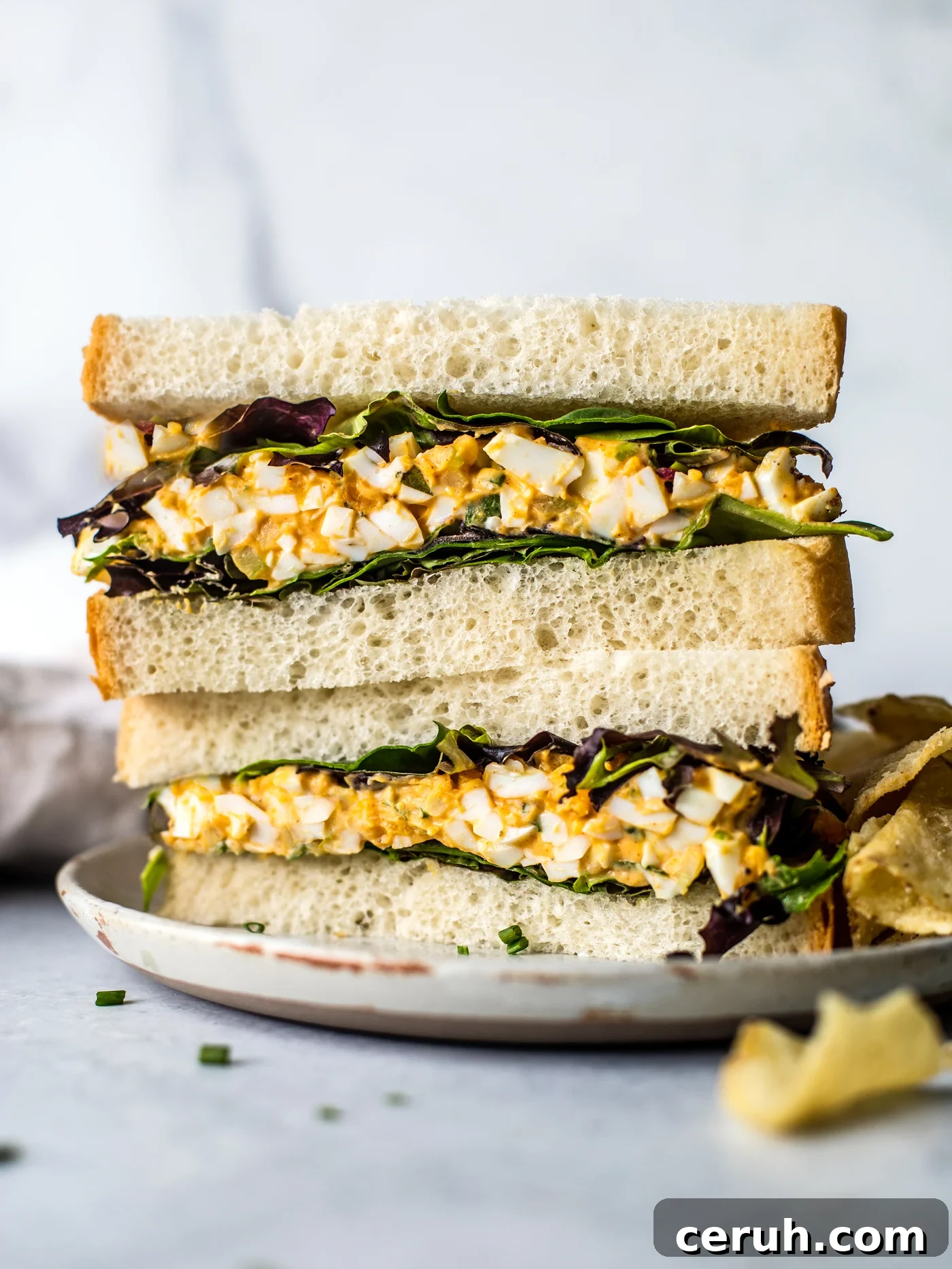 Egg salad sandwich halves with mixed greens stacked on a plate with a side fo chips. 