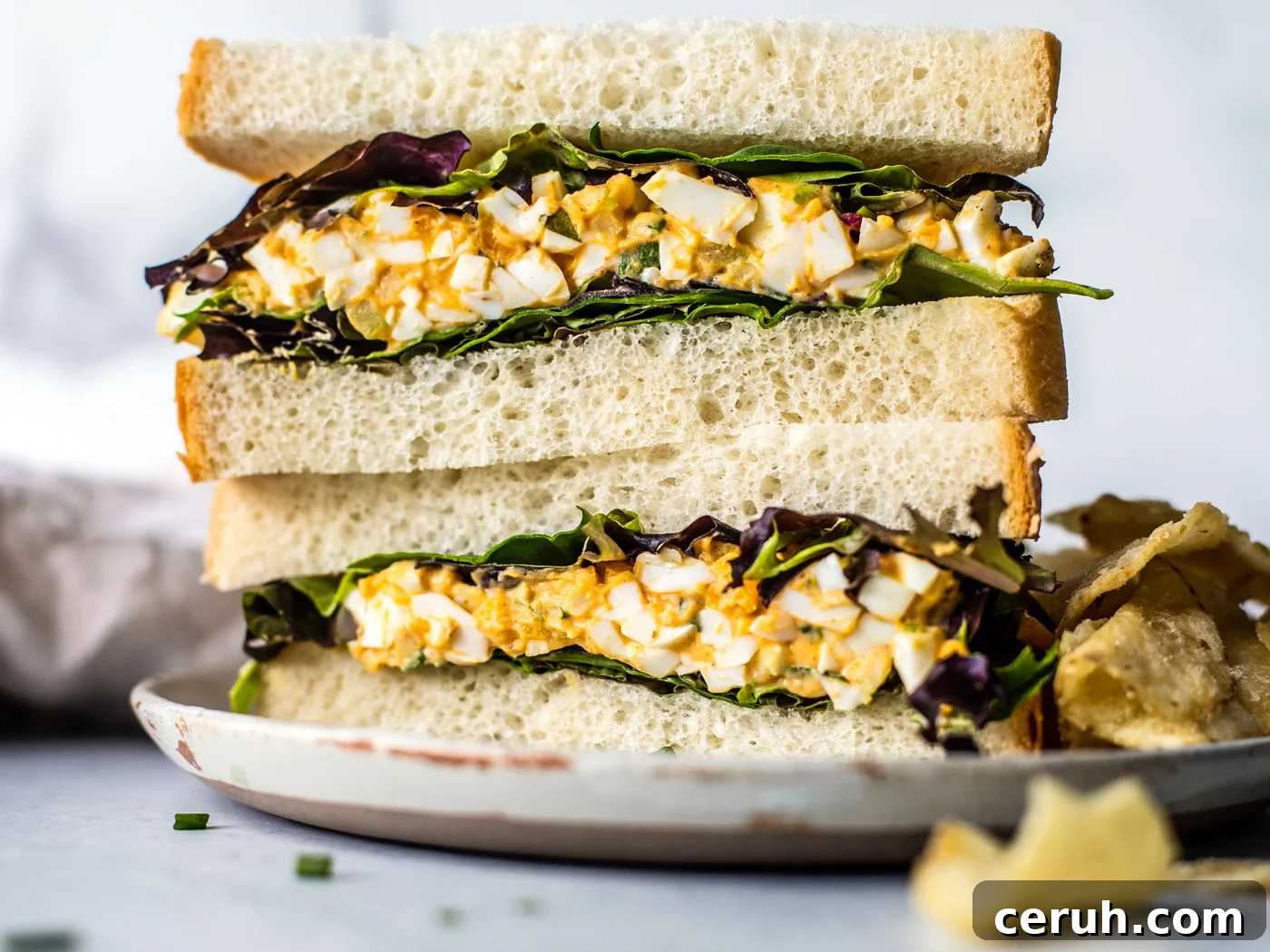Egg salad sandwich halves with mixed greens stacked on a plate with a side of chips.