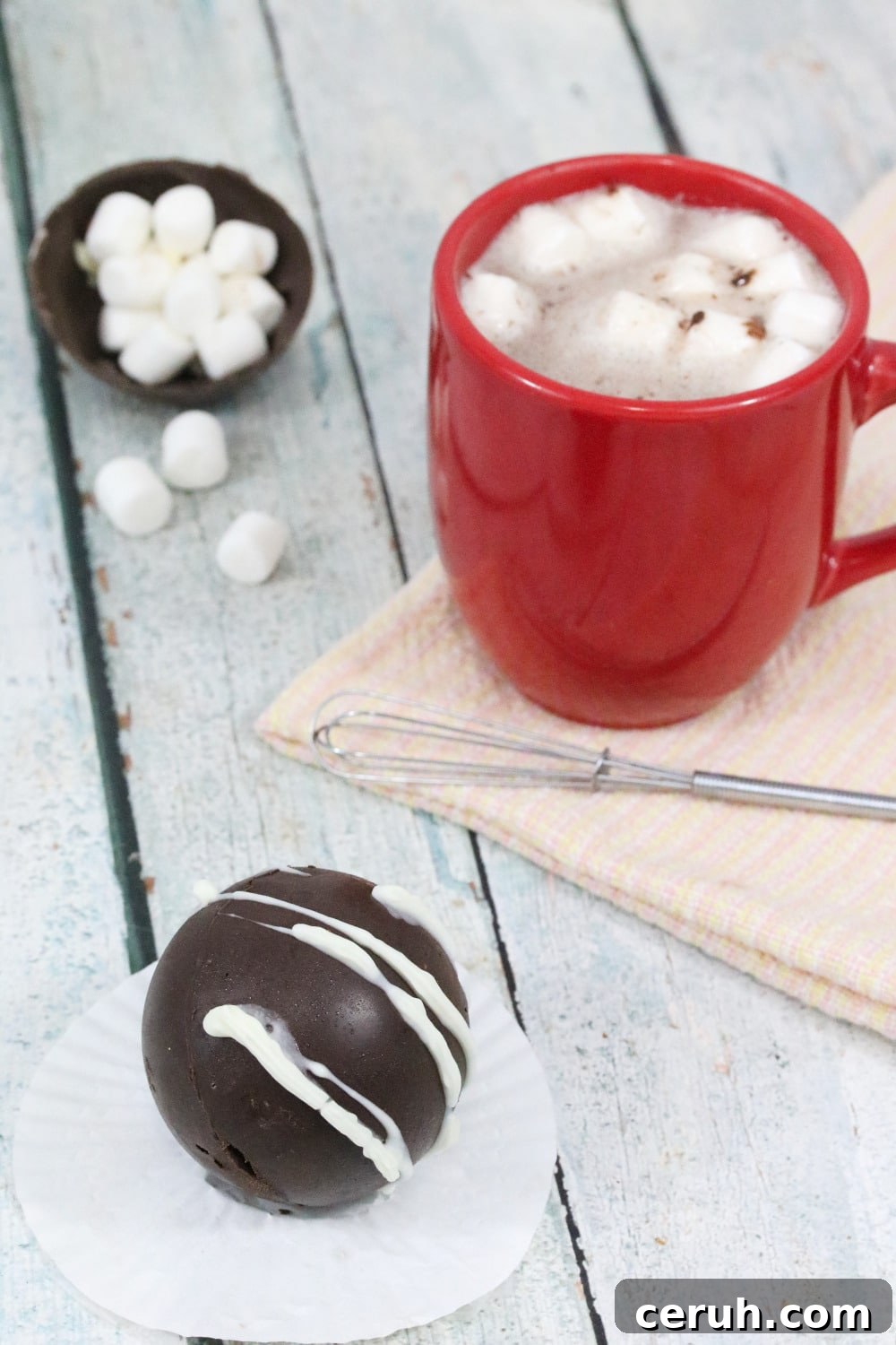 hot chocolate bomb on a white cupcake liner with more bombs in a white bowl behind it and a red mug of hot chocolate with a whisk and marshmallows