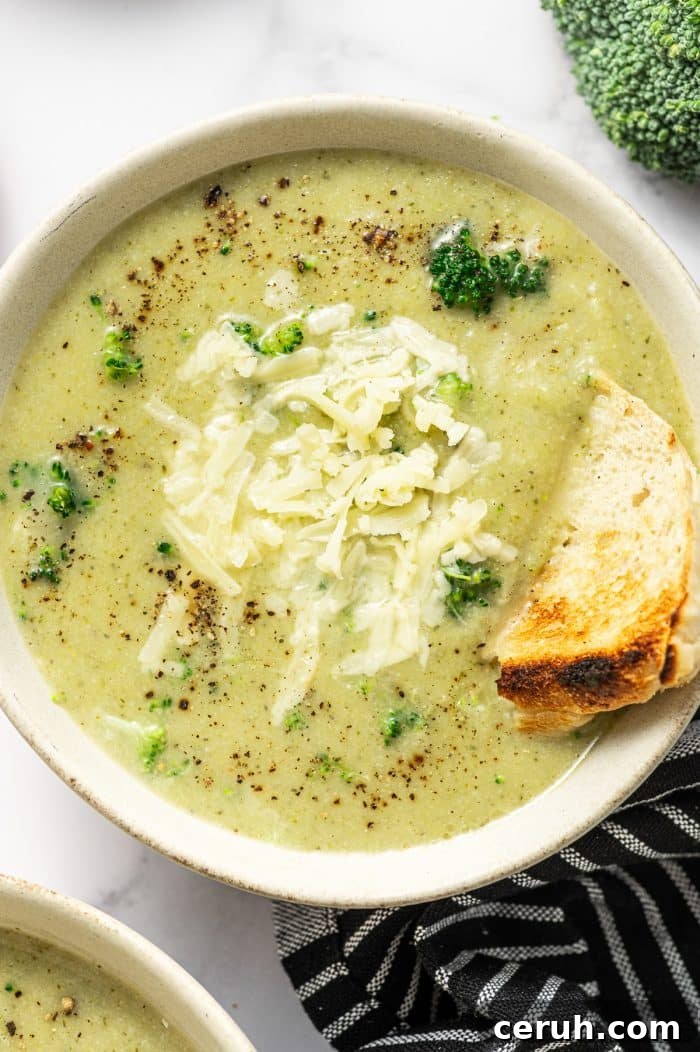 Overhead photo of a light beige bowl filled with creamy pale green broccoli cauliflower soup topped with shredded cheddar and cracked black pepper. A toasted bread wedge is tucked into the right side of the bowl for dipping.