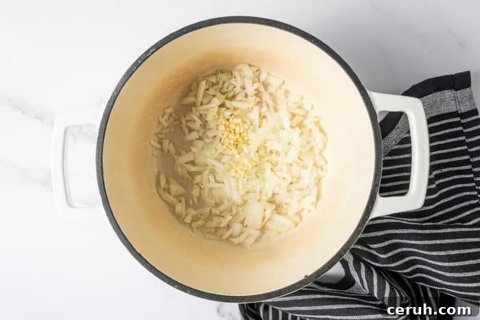Overhead shot of a cream-colored enameled Dutch oven with a black rim and white handles. Chopped onion and minced garlic are sautéing in the bottom of the pot.