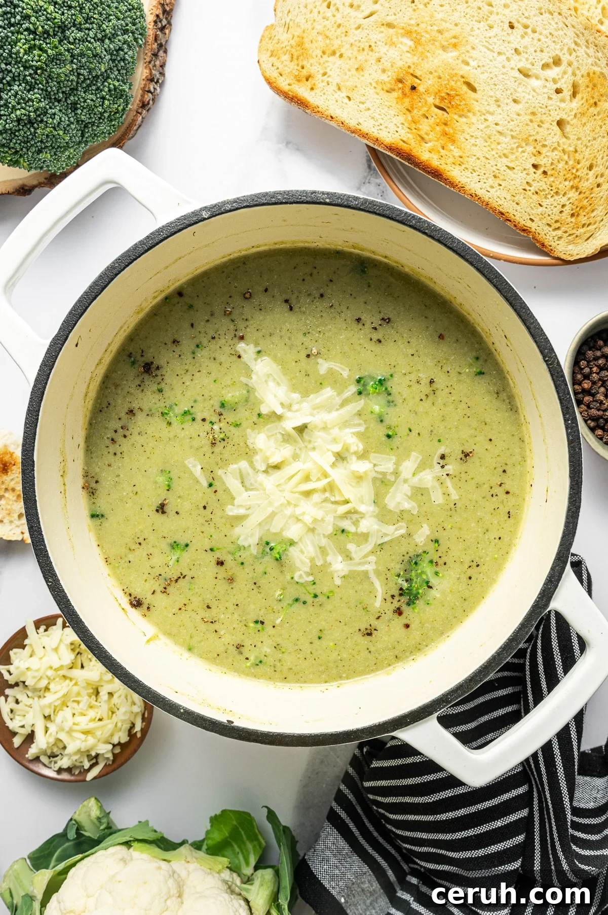 Overhead hero photo of broccoli cauliflower soup in a cream enameled Dutch oven. The soup is creamy pale green and topped with shredded cheddar, scattered black pepper, and a few small broccoli florets.