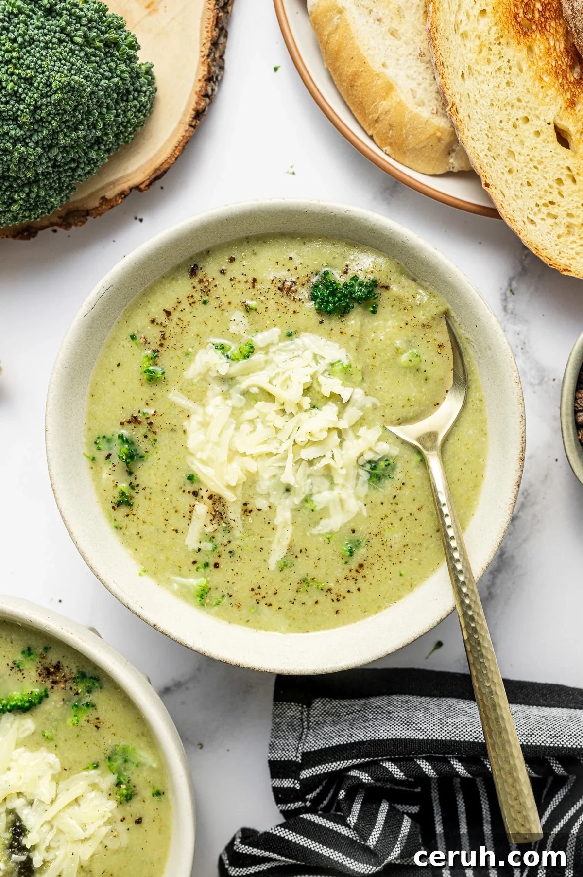 Overhead serving photo of broccoli cauliflower soup in a light beige bowl. The soup is topped with shredded cheddar, cracked black pepper, and a few broccoli bits, with a gold-handled spoon resting in the bowl.