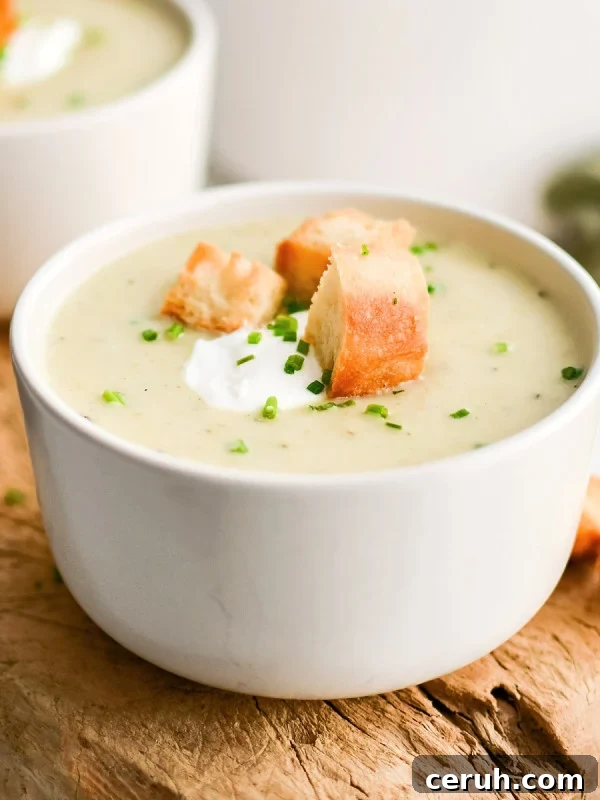 White soup bowl on a rustic wood board filled with homemade creamy potato leek soup and garnished with sour cream, homemade croutons, and freshly chopped chives.