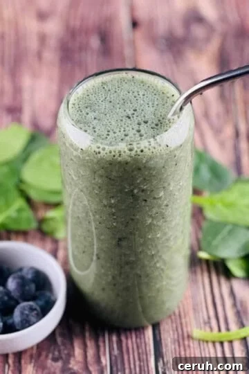 A delicious spinach smoothie poured into a tall glass, with a small bowl of blueberries beside it, and spinach leaves behind it.