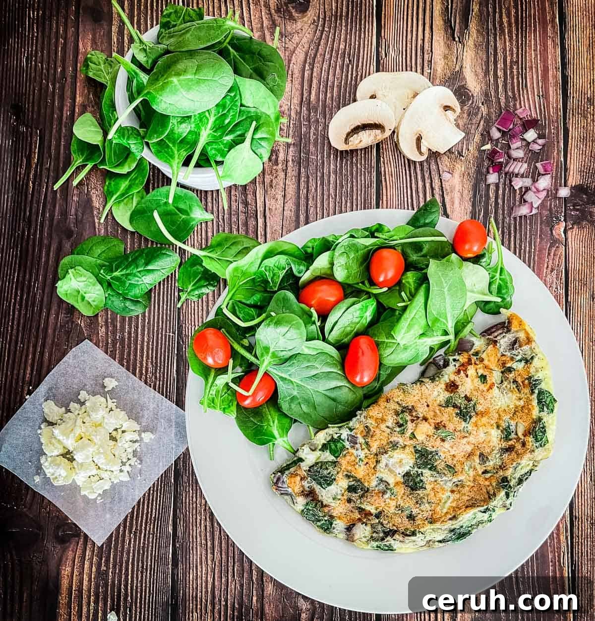 The spinach omelette on a white plate with a side salad with the ingredients to make the omelette beside the plate.