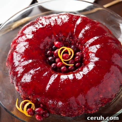 red cranberry jello salad in a ring on a glass cake stand with orange peel and fresh cranberries as garnish.