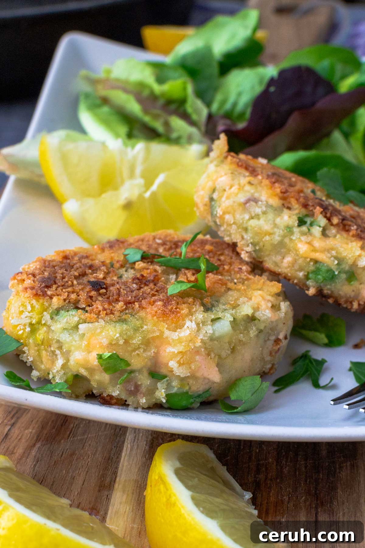 Closeup of 2 crispy salmon fish cakes on a rectangular white plate with green salad and lemon segments.