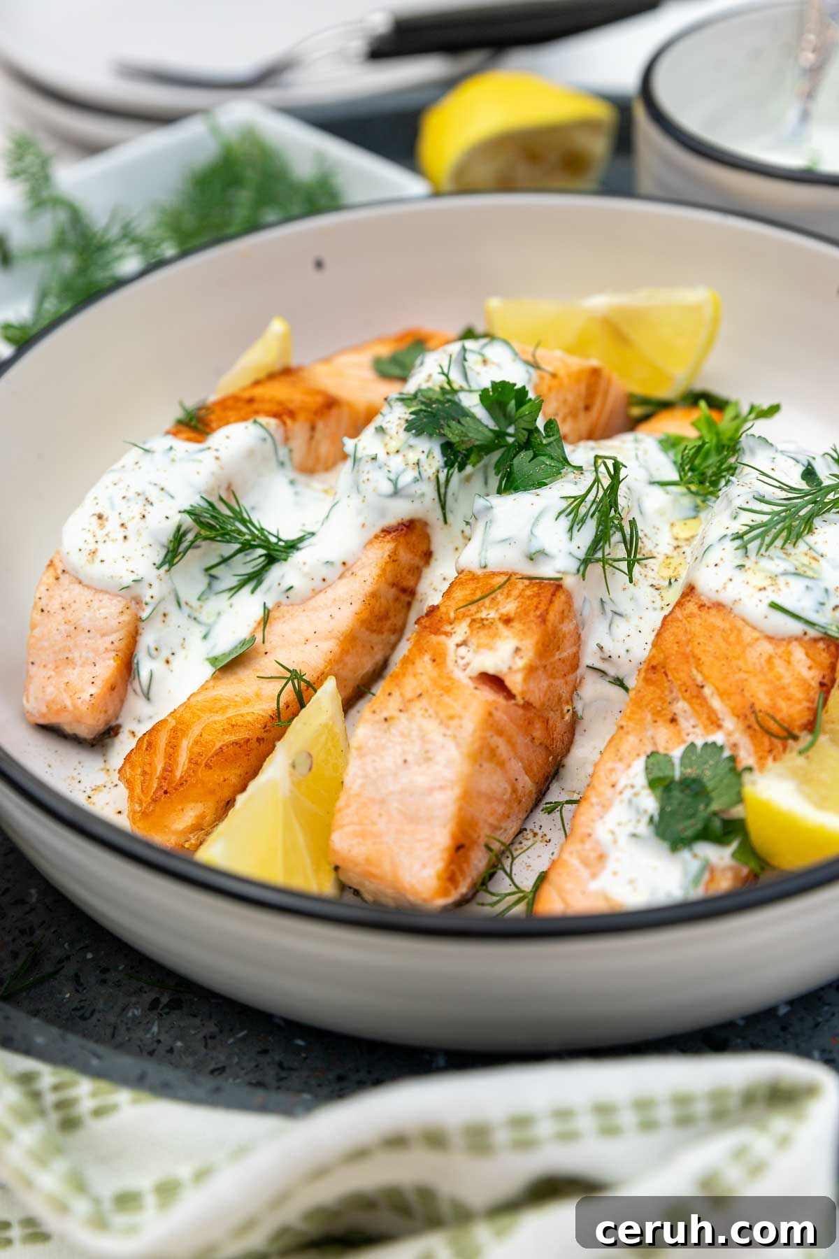 Closeup of a dish of cooked salmon with dill yogurt sauce drizzled over it garnished with lemon segments and herbs.
