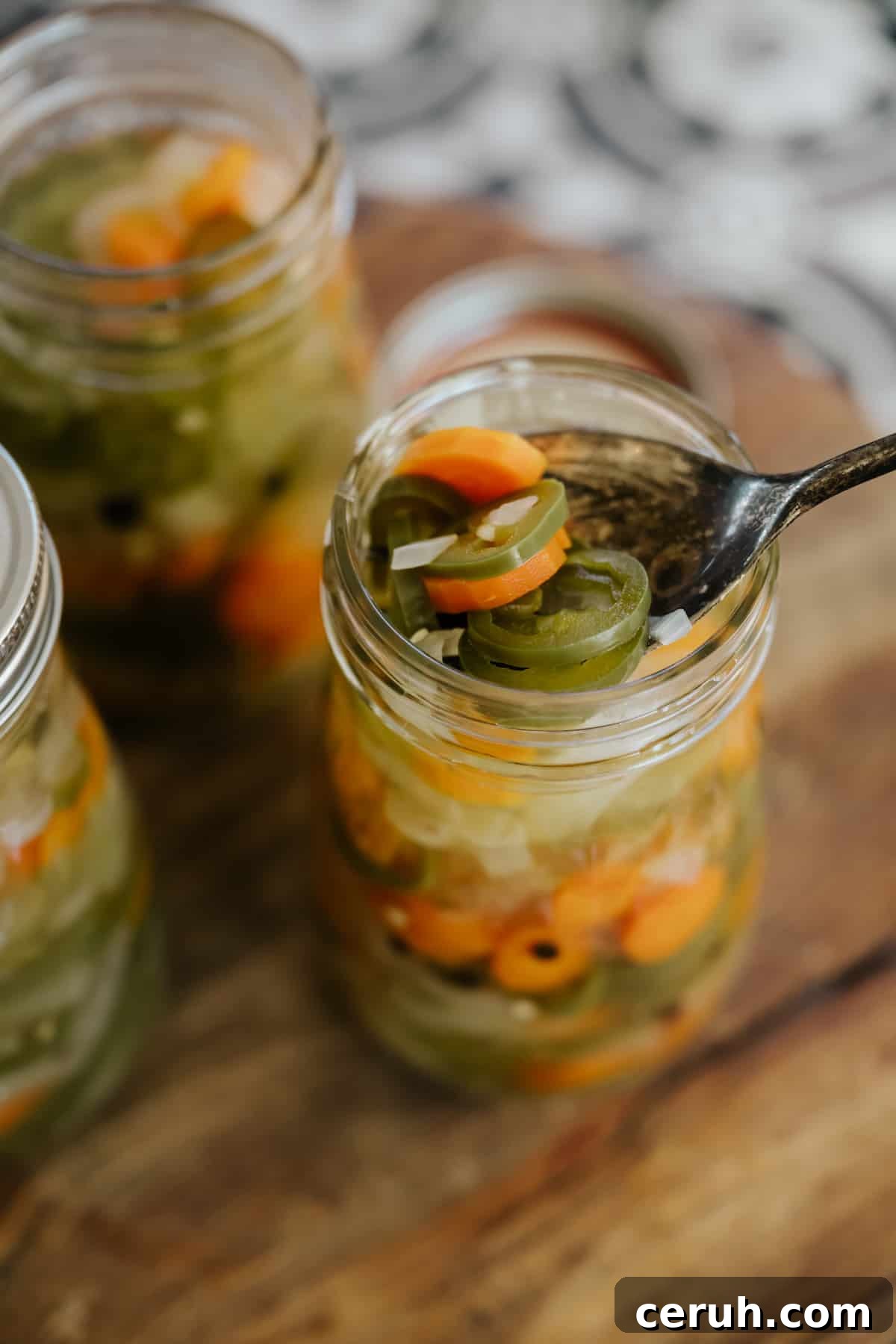 multiple mason jars filled with escabeche pickled jalapeños in glass jars on a wooden cutting board with the closest one with a spoon in it fishing out a serving.