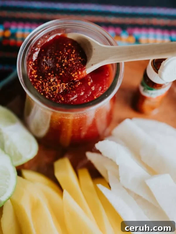 Homemade chamoy recipe by Yvette Marquez-Sharpnack of Muy Bueno, a sweet, sour, spicy Mexican chamoy sauce served in a jar with a wooden spoon, topped with chile lime seasoning and paired with fresh mango and jicama.