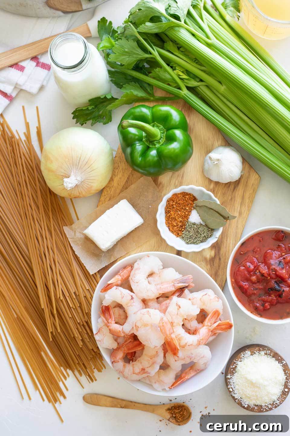 Overhead of ingredients, including raw shrimp in bowl, Cajun spices, dry pasta, and uncut veggies gathered on wooden cutting board.